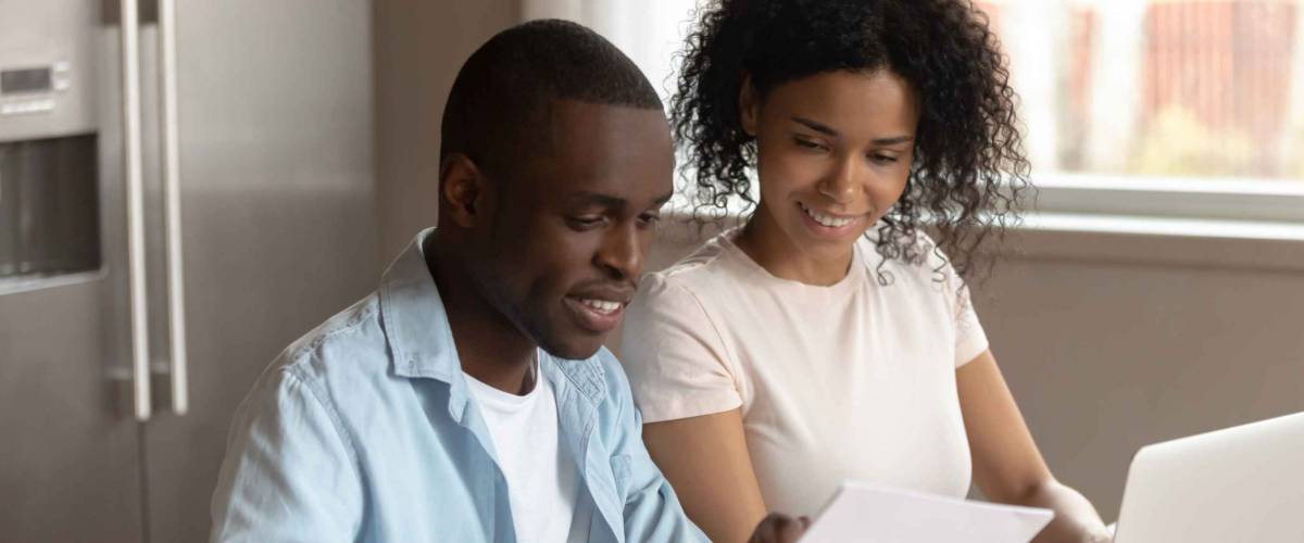 Smiling couple holding piece of paper, working at laptop at kitchen table