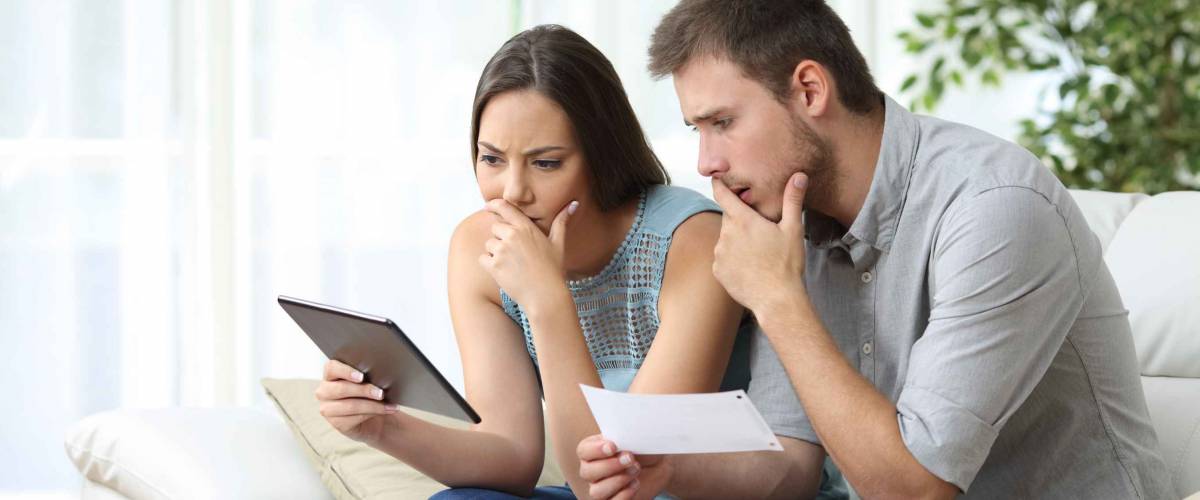 Concerned looking couple sit on couch, looking at tablet and pieces of paper