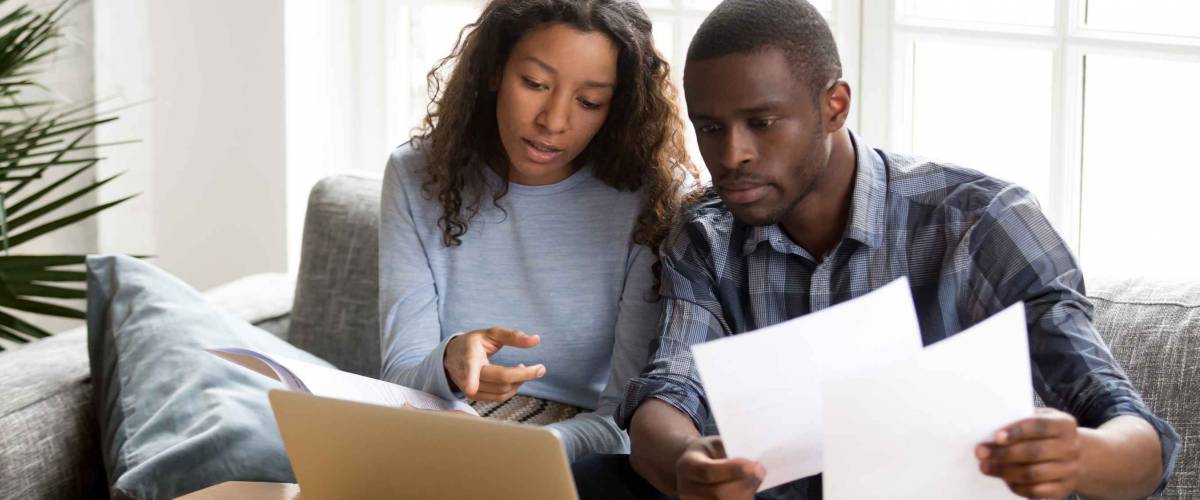 Serious African American couple discussing paper documents, sitting together on couch at home, man and woman checking bills, bank account balance, terms of contract, mortgage, loan agreement
