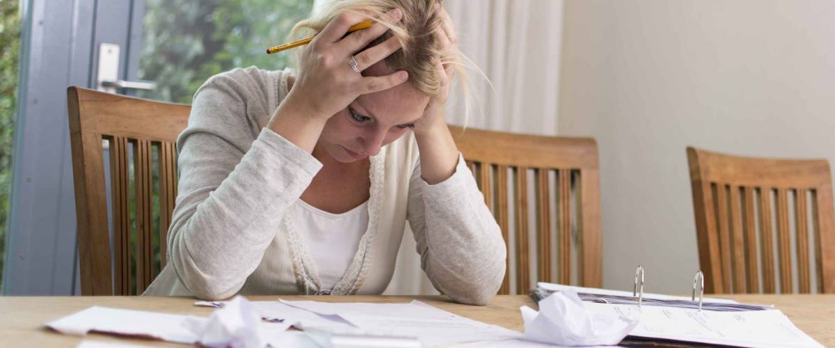Woman holding head in hands, looking down at pile of bills on kitchen table