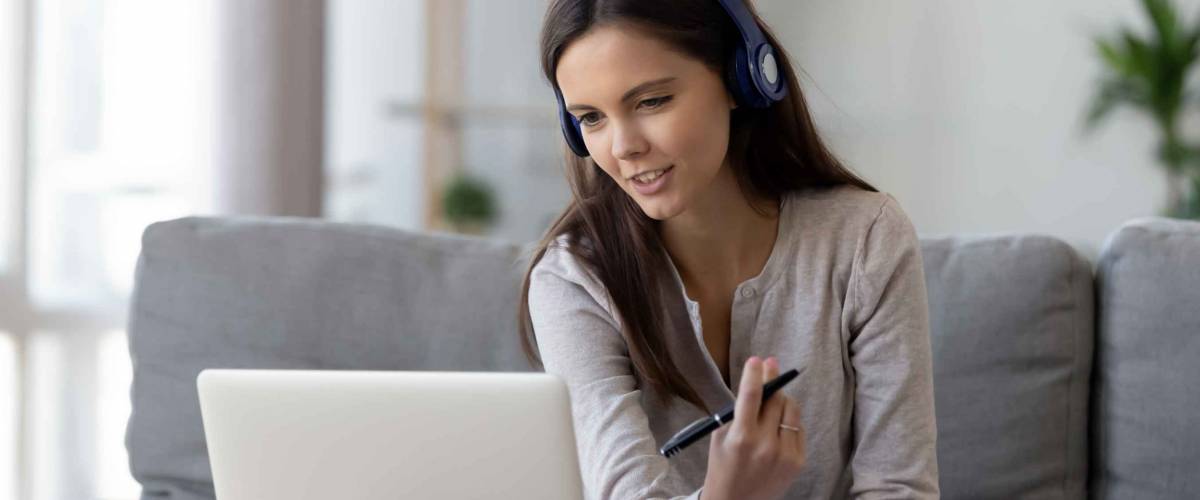 Young woman sits on couch, wearing headphones, talking to laptop and taking notes