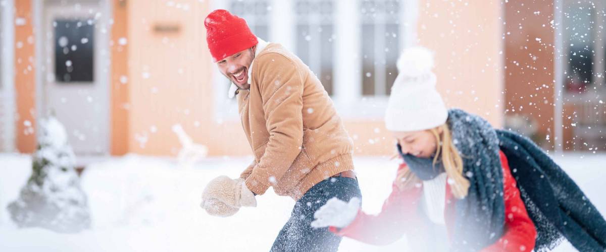 Young Beautiful Couple Throws Snowballs at Each other While Snow Falls. Happy Man and Woman Playing with Snow in the Yard of their Idyllic House. Family Enjoying Winter.