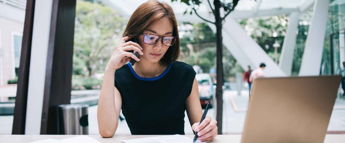 Woman wearing glasses holds cellphone to her ear, writes notes while working on laptop