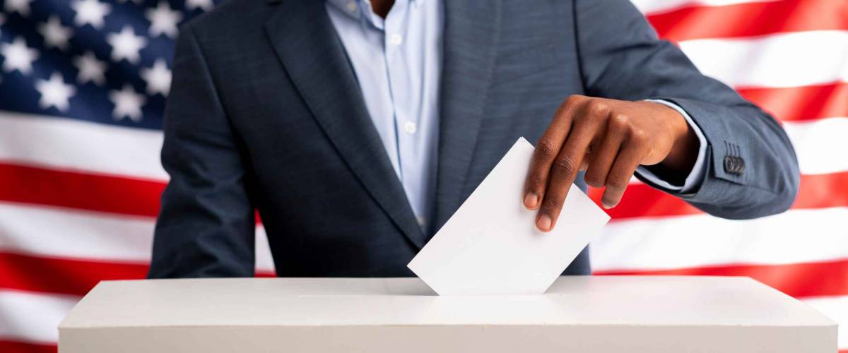 USA elections 2020. Happy african american man voting inserting paper into ballot box over american flag background