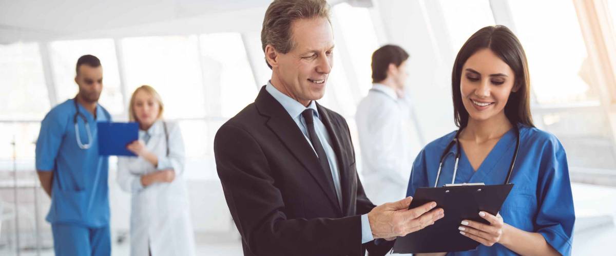 Handsome mature businessman and beautiful young doctor are discussing documents and smiling while standing in the hospital hall