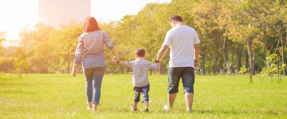 Mother and father holding hands with young child between them, walking in a park in the sun