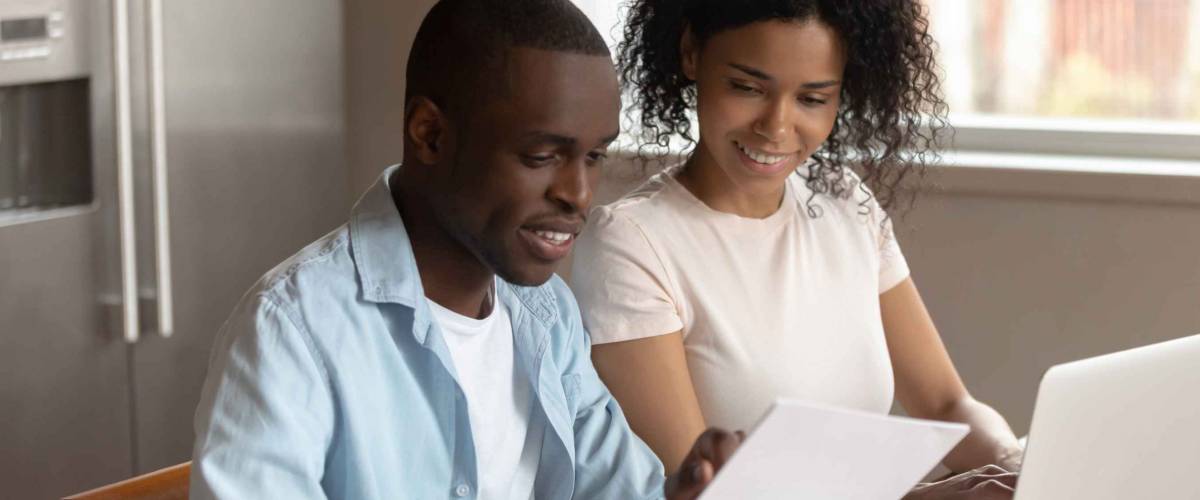 Happy couple sitting at kitchen table, looking at forms and using laptop