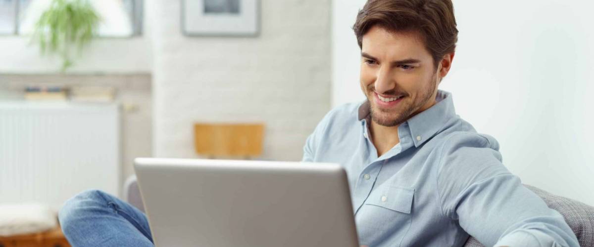 Young man smiling as he reads the screen of a laptop computer while relaxing on a comfortable couch at home in his jeans