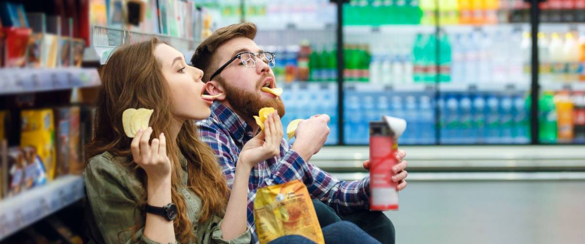 Portrait of a young funny hungry couple sitting on the supermarket floor and eating junk food