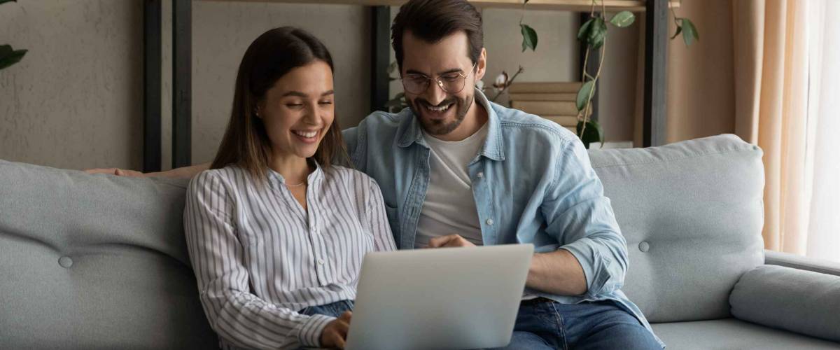 Happy husband wife newlyweds rest on sofa hold laptop.