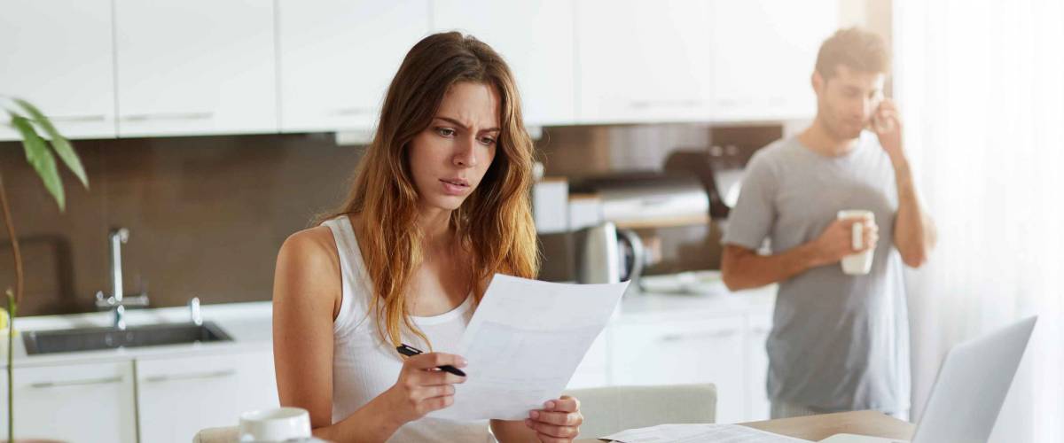 Woman looking at paper sitting at kitchen table, while man in the background on the phone.