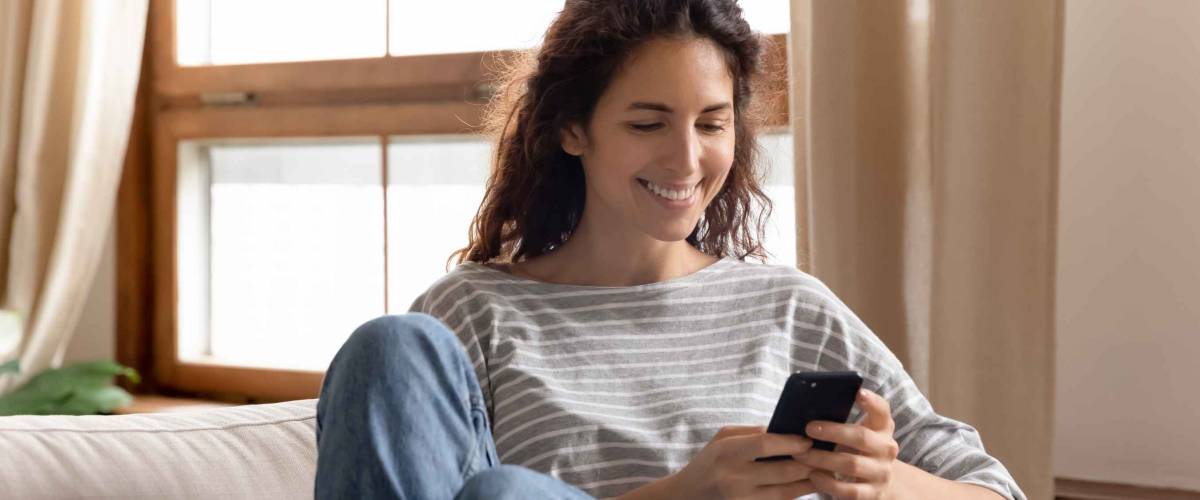 Happy young woman sitting on the couch, using her smartphone
