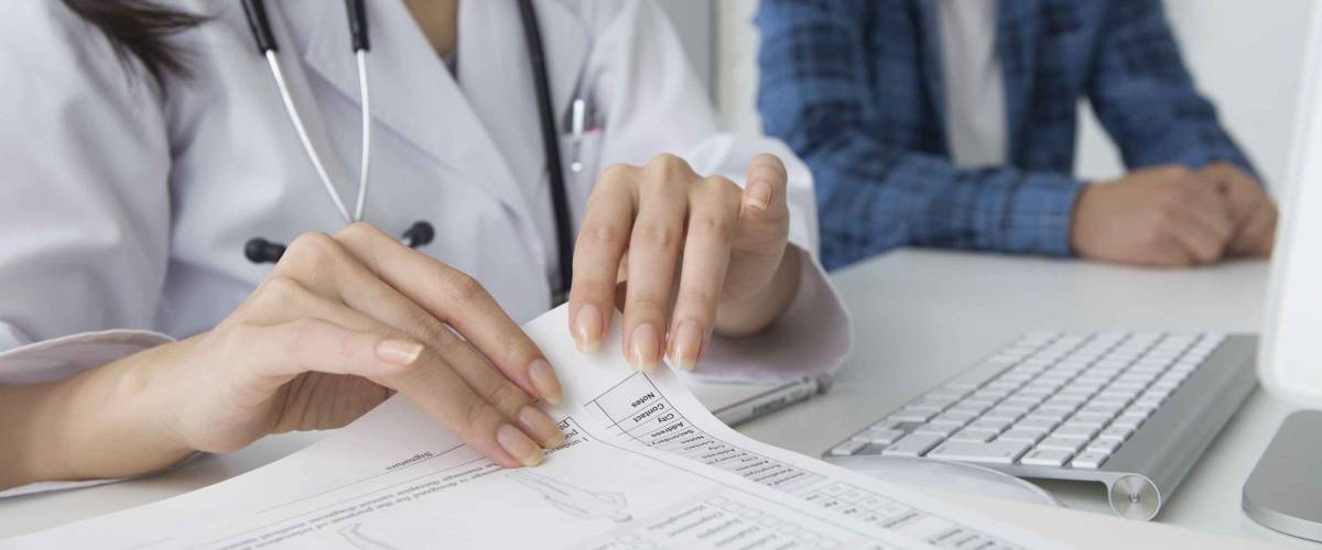 Doctor holds papers on table in foreground, man sitting in the background