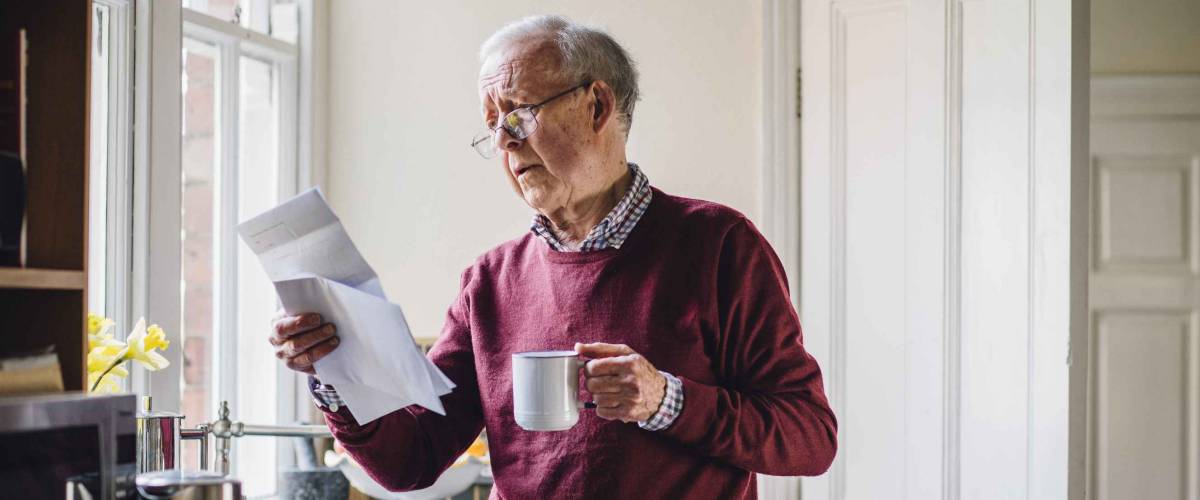 Senior man is standing in the kitchen of his home with bills in one hand and a cup of tea in the other. He has a worried expression on his face.