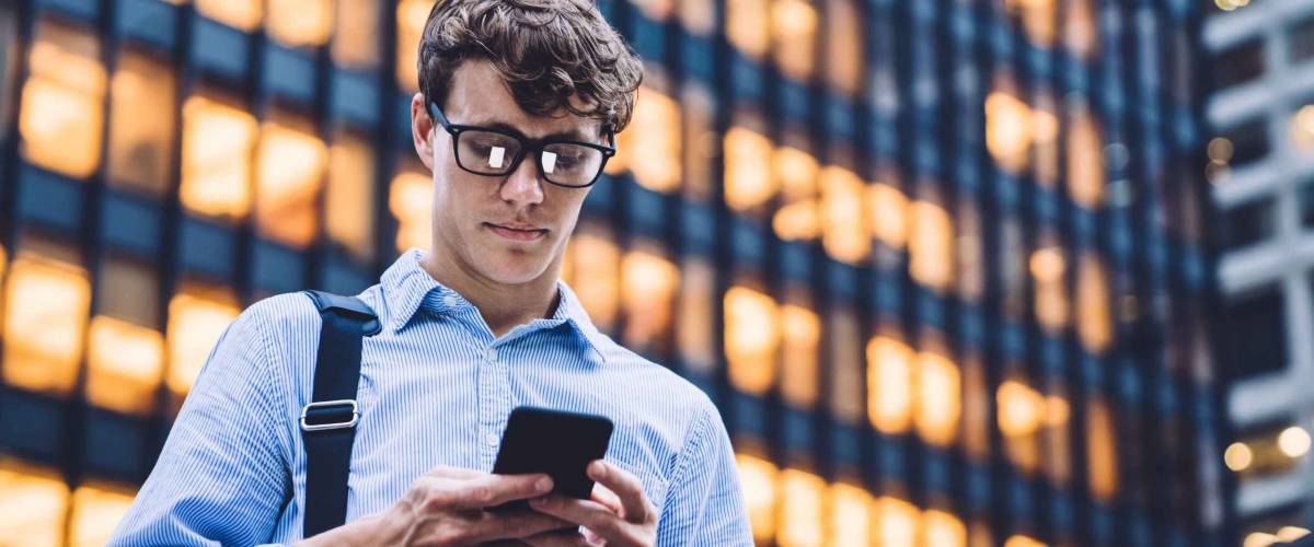 Man in shirt with rolled sleeves and glasses with bag texting on phone standing on background of lighted high building