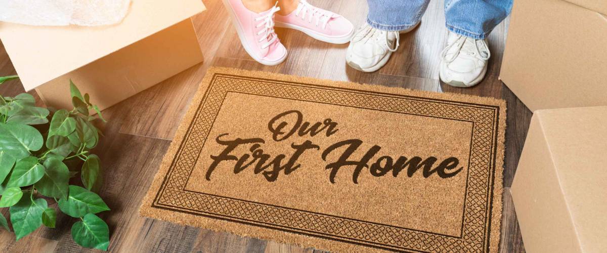Man and Woman Unpacking Near Our First Home Welcome Mat, Moving Boxes and Plant.
