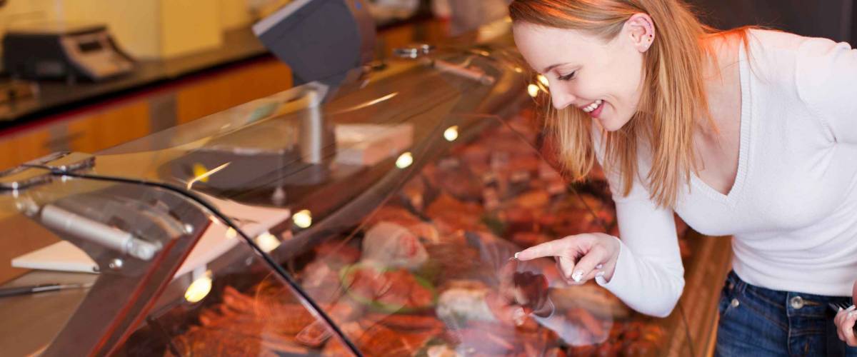 Happy young woman choosing meat from glass cabinet in grocery store