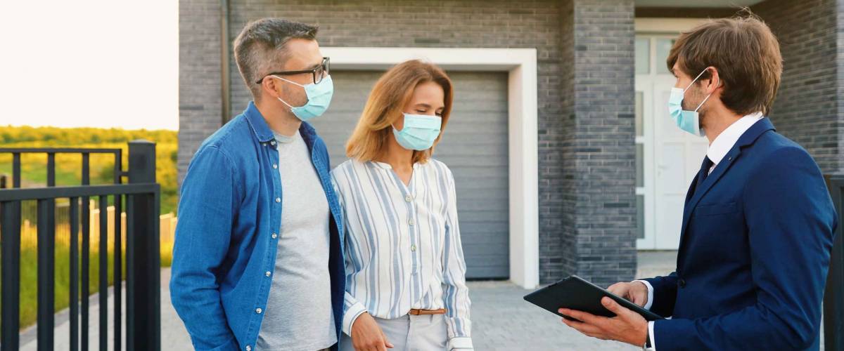 Caucasian young happy couple in medical masks buying house at outskirt and talking with male real-estate agent holding tablet device in hands. Outdoor. Coronavirus pandemic concept.