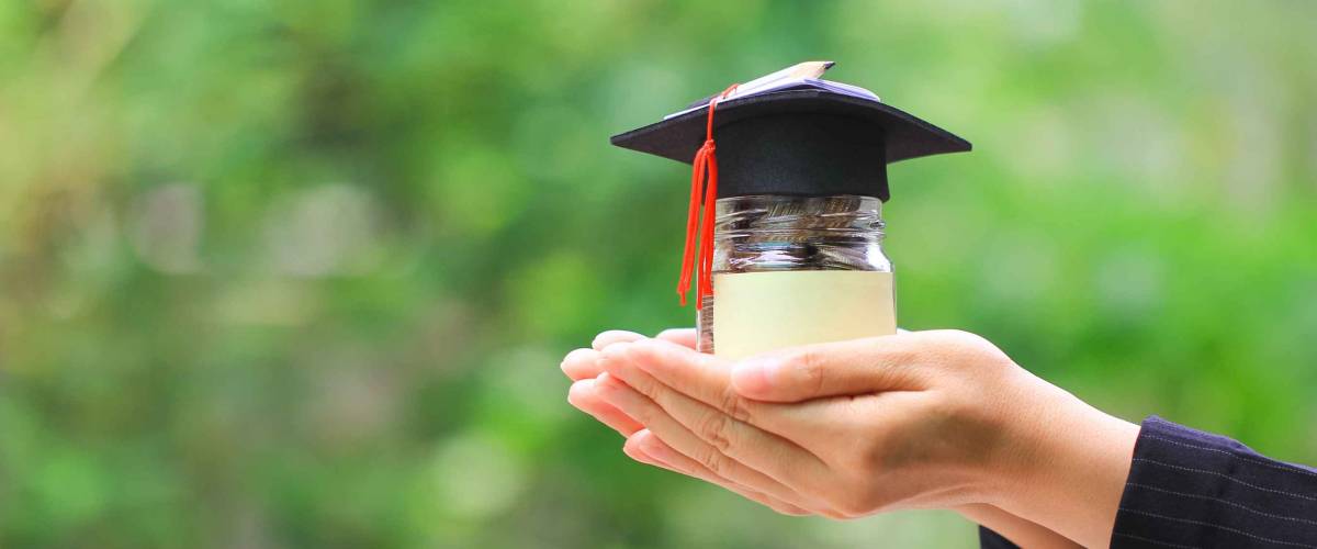 Woman hand holding coins money in glass bottle with graduates hat on natural green background, Saving money for education concept