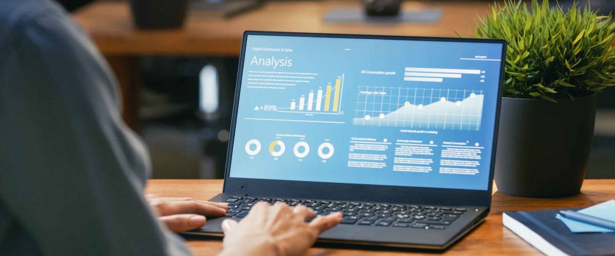 Female Analyst at Her Desk Works on a Laptop Showing Statistics, Graphs and Charts. She Works on the Wooden Table in Creative Office. Over the Shoulder Footage.