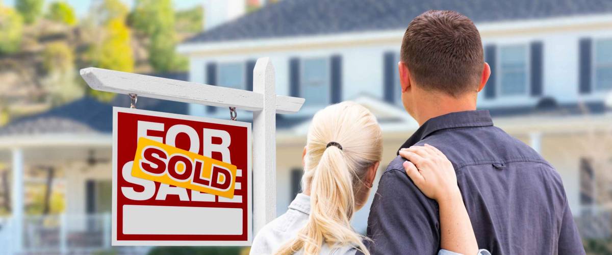 Young Adult Couple Facing Front of Sold Real Estate Sign and House.