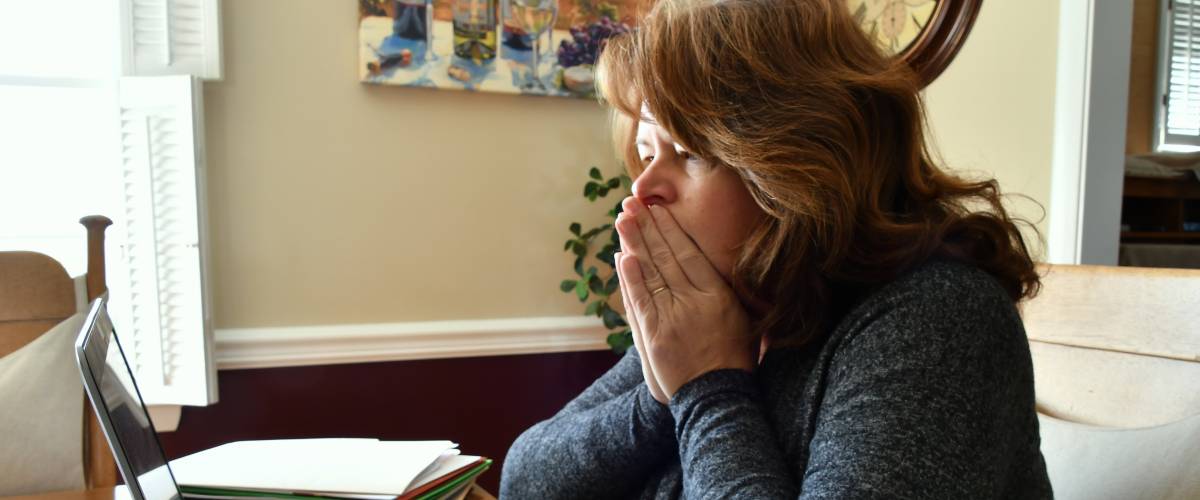 Woman sits in front of computer, holding hands in front of face, looking upset