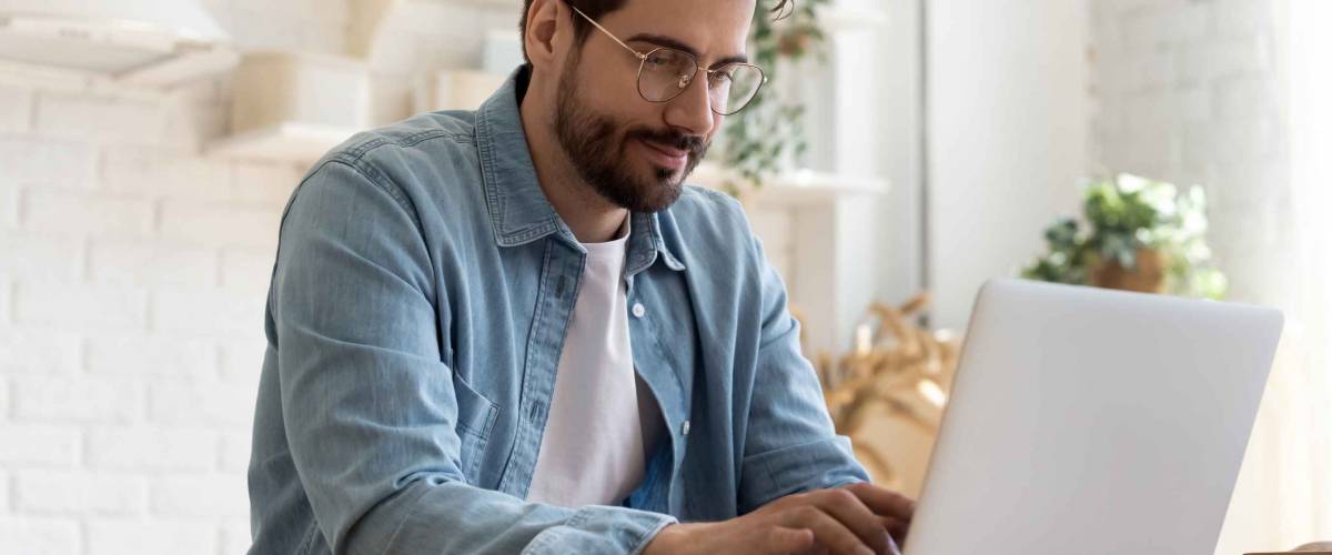 Focused young man wearing glasses using laptop, typing on keyboard, writing email or message, chatting, shopping, successful freelancer working online on computer, sitting in modern kitchen