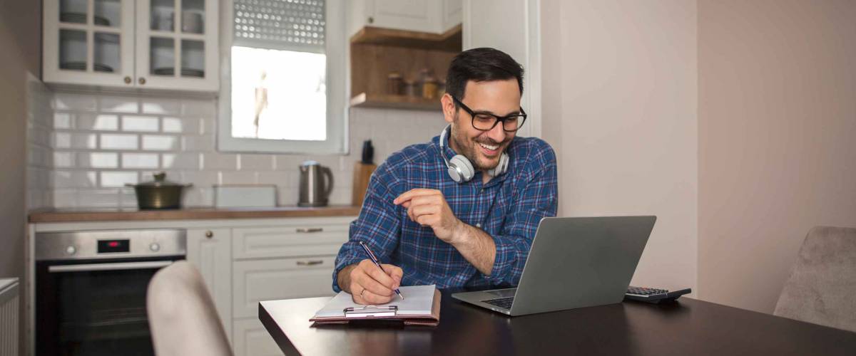 Young smiling businessman working from home writing notes in notebook while using his laptop and wearing headphones around his neck.