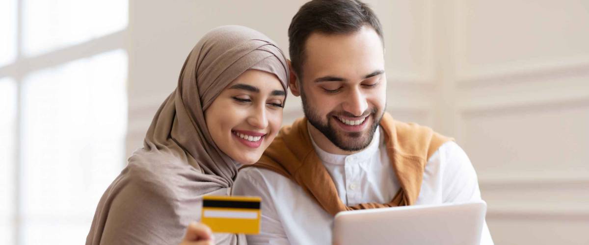 Online Shopping. Happy Muslim Couple Using Digital Tablet And Credit Card Buying And Purchasing Things Sitting On Sofa At Home. Joyful Customers, Modern Shopping And Sales Concept