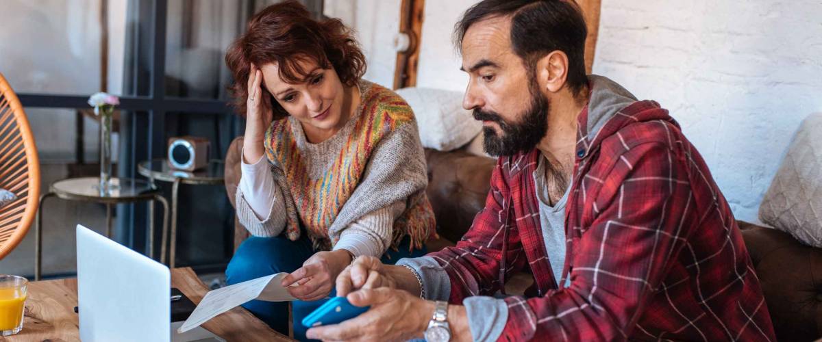 Couple sitting on couch, looking distressed while looking at papers, phone and computer.