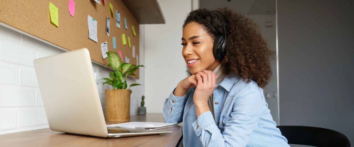 Smiling young woman sits at desk, wearing headphones and looking at laptop.