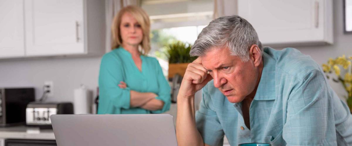Older senior married couple in distress, looking at laptop on the kitchen counter.