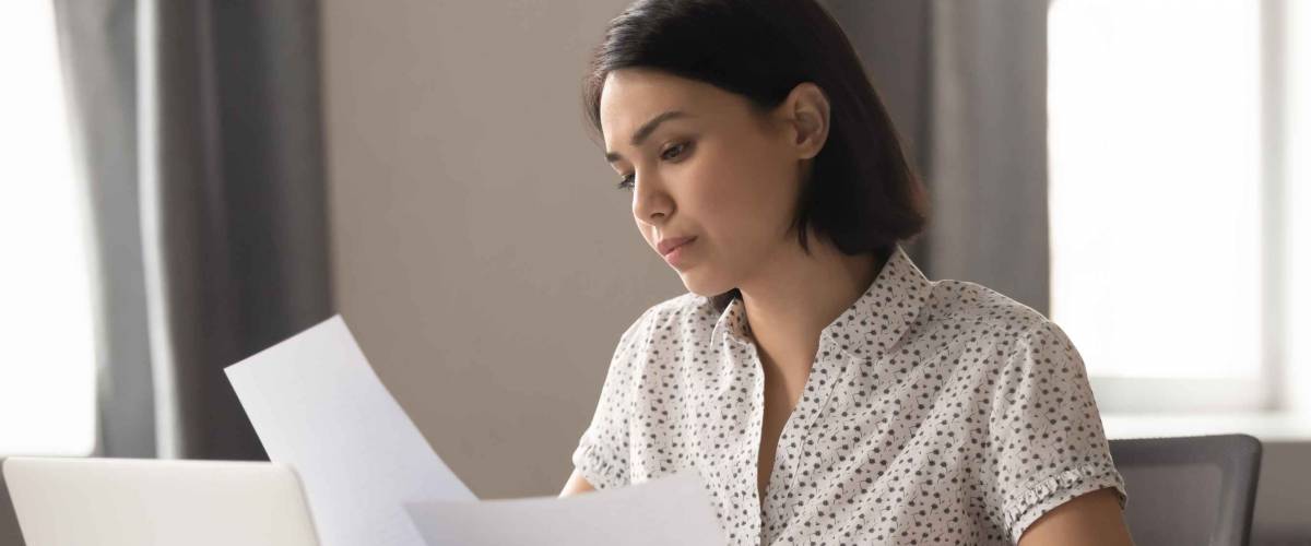 Woman holding pieces of paper, looking seriously down at them sitting at a desk.