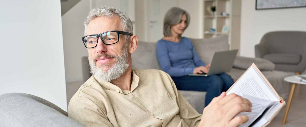 Happy older senior man husband wearing glasses reading book relaxing sitting on couch at home with mature wife using laptop. Middle aged family couple enjoying casual daily activities