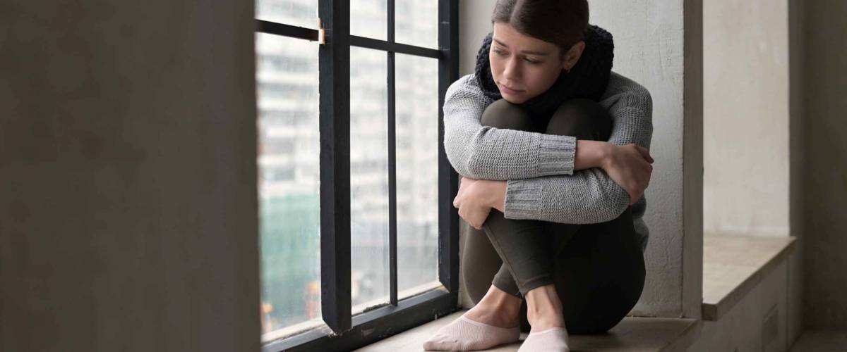 A sickly-looking young woman is sad on the windowsill of a city apartment. Autumn depression of a city dweller