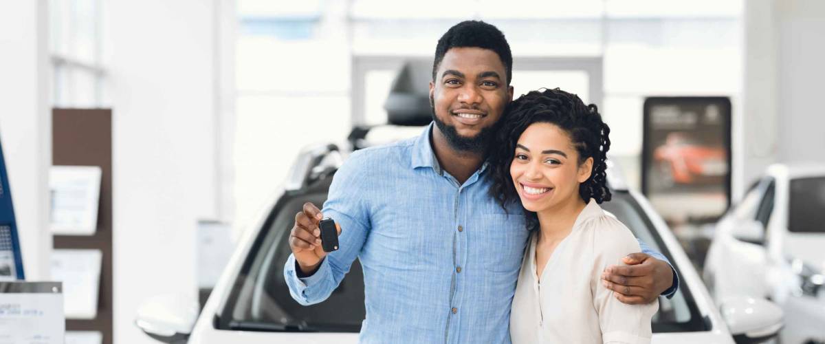 Happy-looking couple standing holding a key in front of a white car at a dealership.