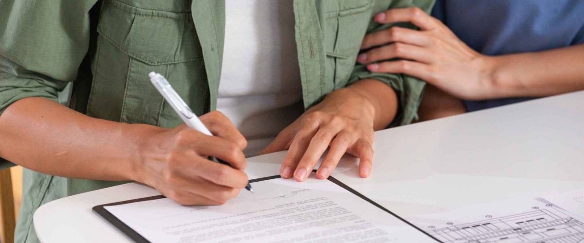 Close up of one woman holding another woman's arm as she signs a contract.
