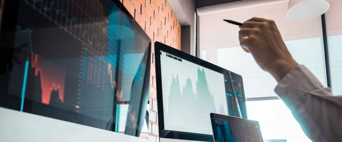 Man's hand points at graphs on computer screens.