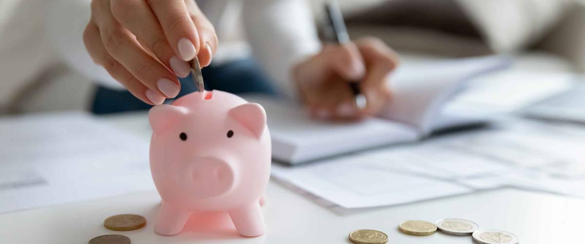 Close up of young woman dropping coins in a pink piggybank while writing notes in a notebook.