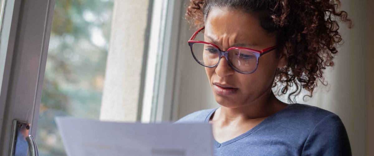 Woman sitting near window squinting down looking at a piece of paper, upset.