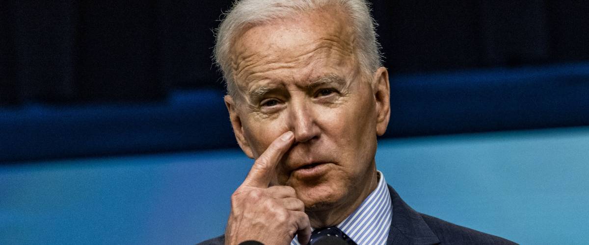Biden stands at a podium and scratches his nose with one finger.