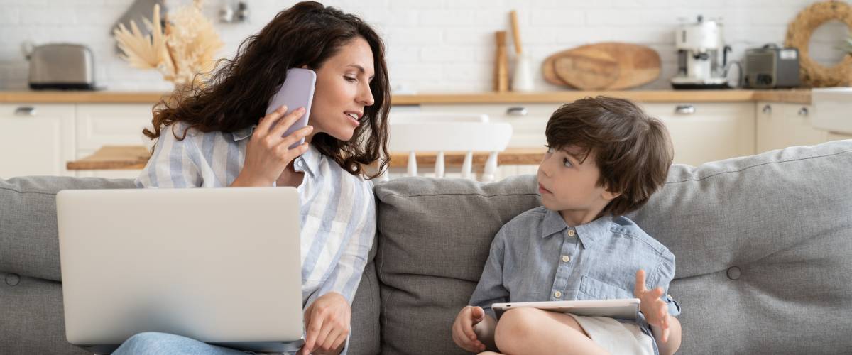 Woman sits on the couch on the phone, next to a little boy showing her something on a tablet.