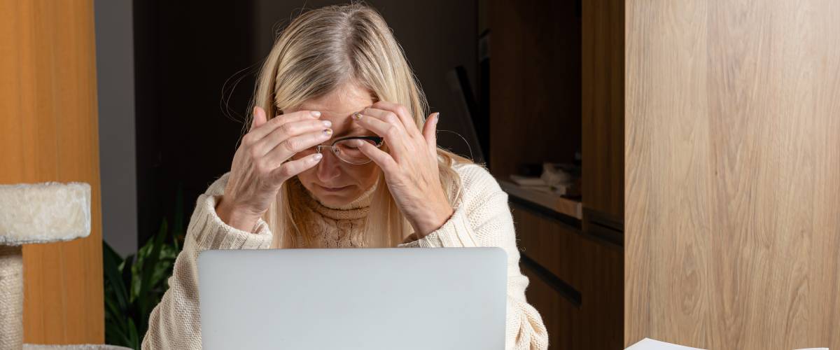 Middle-aged woman sits at desk, with head in her hands, looking distressed.