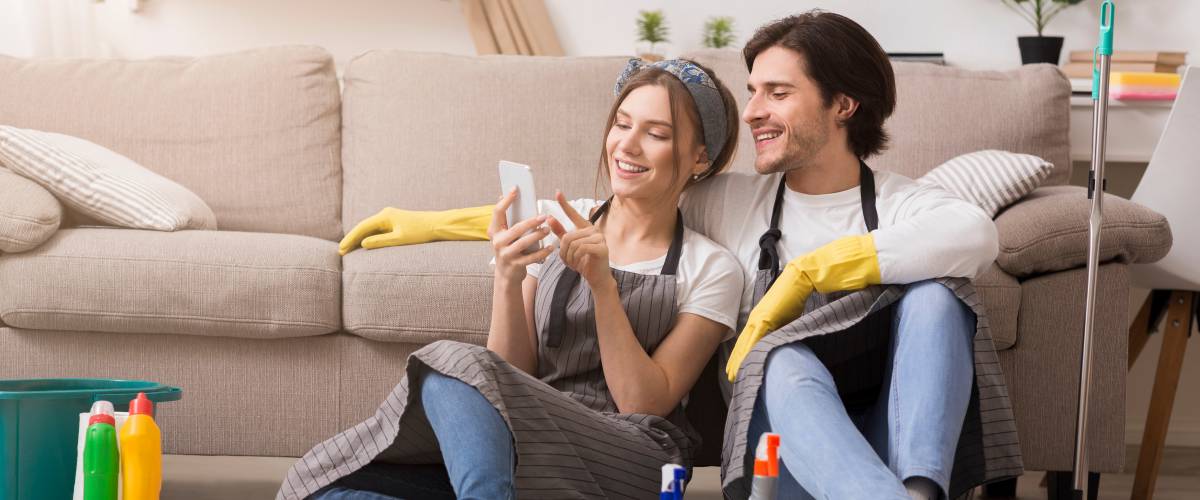 Smiling young couple relaxing with smartphone after spring-clean apartment, sitting on floor with lots of detergents and cleaning supplies, free space