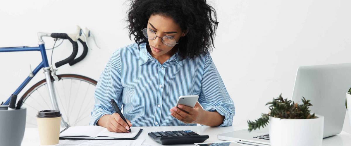Concentrated focused Afro American female freelancer holding phone in one hand and making notes with pen in other while planning budget.