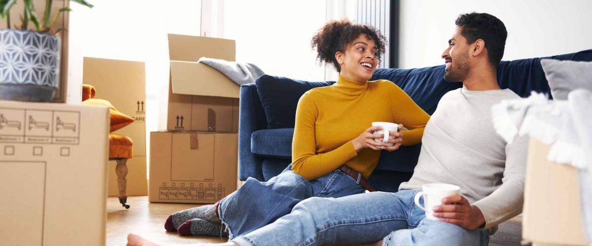 Young mixed ethnicity couple taking a break on moving day into new home sitting on floor in lounge drinking coffee surrounded by removal boxes