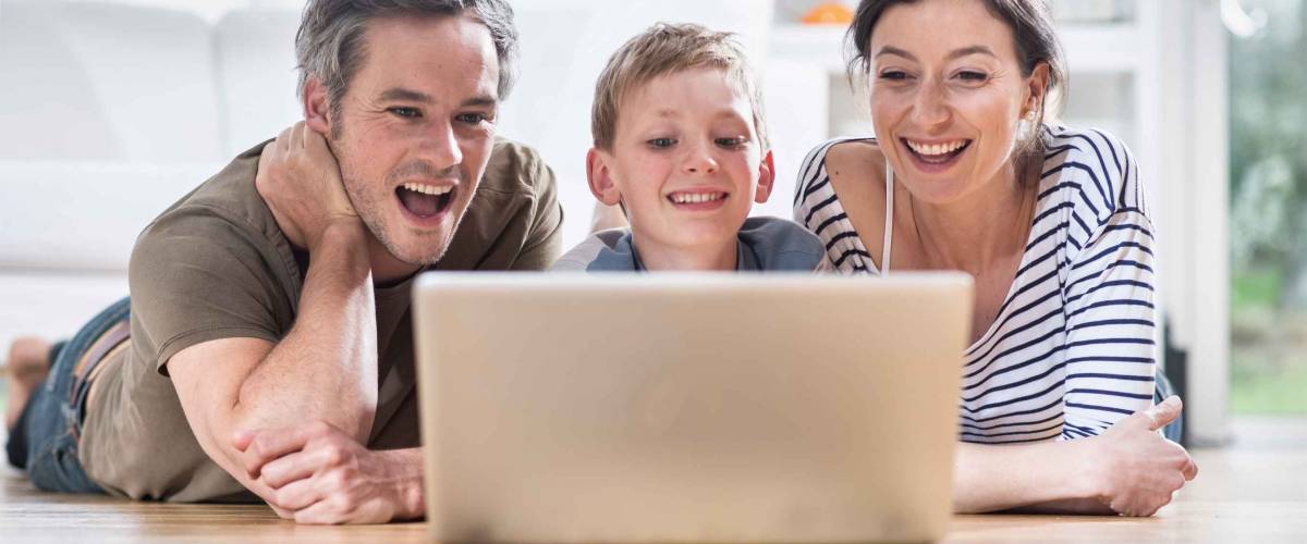 At home, cheerful family sharing a funny video on a laptop. They are lying on the wooden floor of the living room.