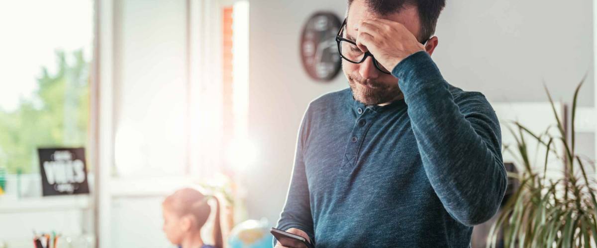 Worried father looking at smart phone and holding hand on forehead, in background his daughter doing homework