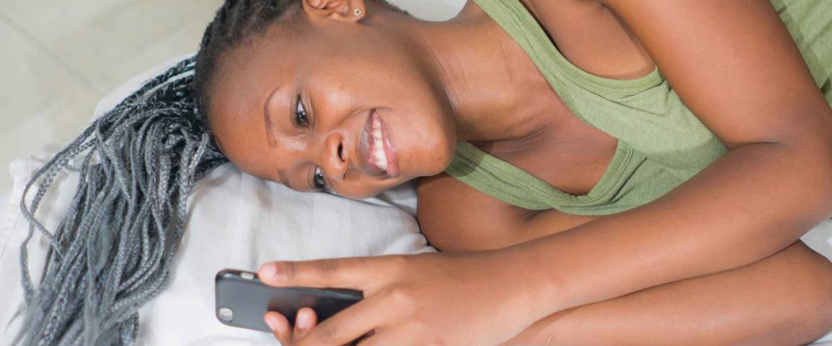 lifestyle home portrait of young happy and attractive black African American woman lying on bed using social media app in mobile phone relaxed and cheerful online dating