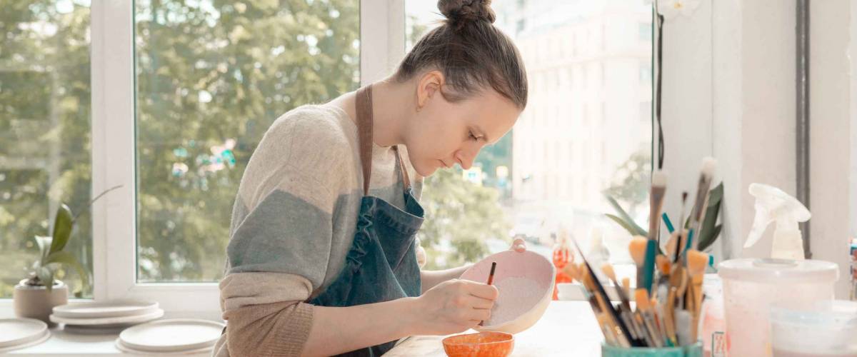 Young woman in apron sitting at table and drawing on ceramic bowl.
